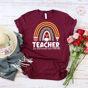 Pu&ograve; includere: Una maglietta color borgogna con un design a arcobaleno e il testo "Teacher all-day-every-day mode". L'arcobaleno &egrave; fatto di matite e ha un cappello di laurea e un cuore al centro.