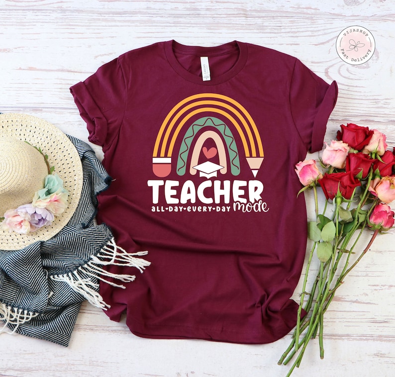 Pu&ograve; includere: Una maglietta color borgogna con un design a arcobaleno e il testo "Teacher all-day-every-day mode". L'arcobaleno &egrave; fatto di matite e ha un cappello di laurea e un cuore al centro.