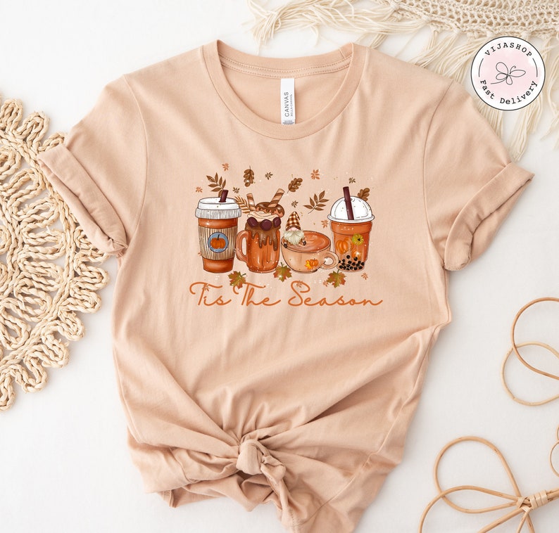 May include: A light peach-colored t-shirt featuring a graphic of four fall-themed beverages, including a pumpkin spice latte and boba tea. The phrase "Tis The Season" is written in a cursive font below the drinks. The shirt is knotted at the bottom.
