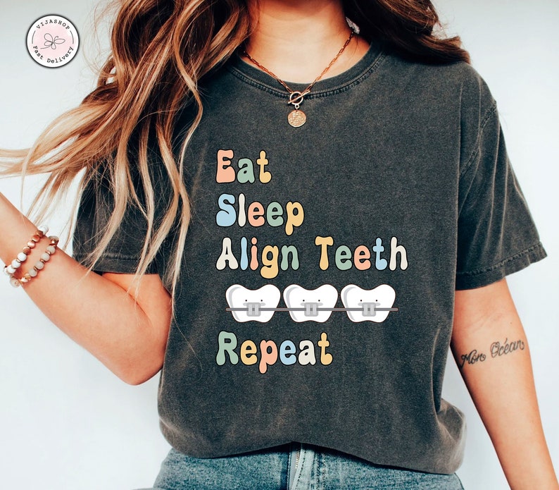 K&ouml;nnte beinhalten: Ein dunkelgraues T-Shirt mit dem Text "Eat Sleep Align Teeth Repeat" in bunten Buchstaben. Der Text ist von drei wei&szlig;en Z&auml;hnen mit Zahnspangen umgeben.