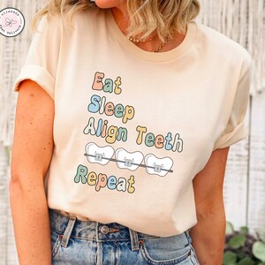 K&ouml;nnte beinhalten: Ein beiges T-Shirt mit dem Text "Eat Sleep Align Teeth Repeat" in bunten Buchstaben. Der Text ist von drei Cartoon-Z&auml;hnen mit Zahnspangen umgeben.