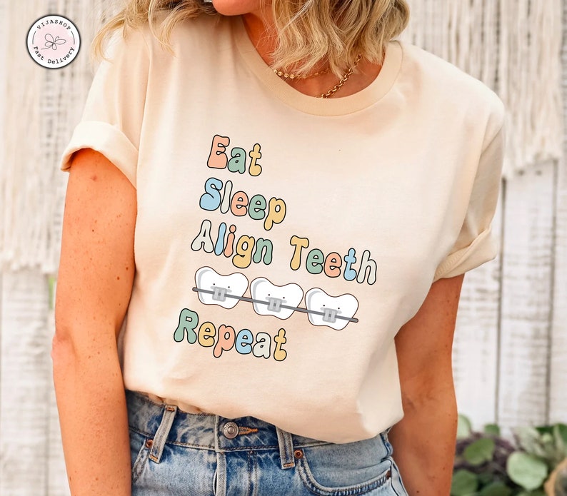 K&ouml;nnte beinhalten: Ein beiges T-Shirt mit dem Text "Eat Sleep Align Teeth Repeat" in bunten Buchstaben. Der Text ist von drei Cartoon-Z&auml;hnen mit Zahnspangen umgeben.