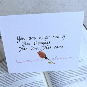May include: A white card with pink calligraphy that reads "You are never out of His thoughts, His love, His care. never alone." A dried pink rose is placed on the card.