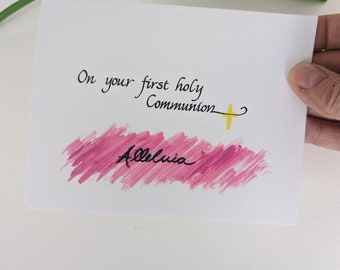 May include: A white card with black calligraphy that reads "On your first holy Communion" with a yellow cross and the word "Alleluia" written in black on a pink watercolor background.