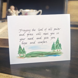 May include: A white card with a handwritten message that reads "Praying the God of all power and grace will meet you in your need and give you hope and comfort." The card has a watercolor illustration of three green trees and a green hill.