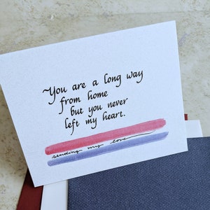 May include: A white card with handwritten text: "You are a long way from home but you never left my heart." Below the text are red and blue brushstrokes with the words "sending my love."