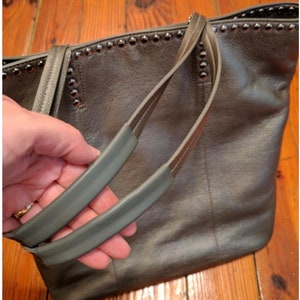 K&ouml;nnte beinhalten: Eine graue Ledertasche mit Nietenbesatz und doppelten Henkeln. Die Tasche hat einen leicht abgenutzten Look.