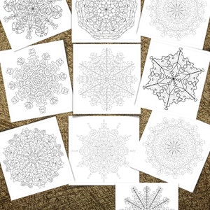 May include: A collection of ten white square coloring pages featuring intricate black and white snowflake designs. A box of colored pencils is visible at the bottom left. The pages are arranged on a textured, golden-brown surface.