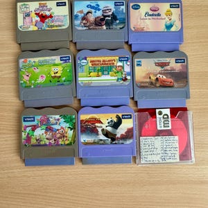 May include: A collection of VTech educational game cartridges featuring various children's characters and movies. The cartridges are in gray and purple plastic cases, with colorful labels. Titles include "Elmo," "Cars," and "Cinderella."