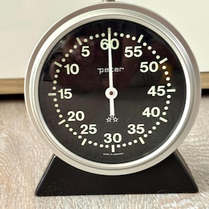 May include: A round analog kitchen timer with a silver metallic frame and a black face. The dial features white numerals from 5 to 60, a single white hand, and the text "peter" and "Made in Germany" printed on the face. It sits on a black base.