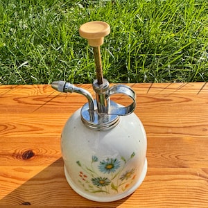May include: A white ceramic plant mister with a floral design and a chrome pump. The mister has a wooden top and a chrome nozzle.