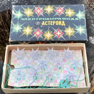 May include: A vintage box of clear star-shaped Christmas lights with a green wire. The box is labelled "Электрогирлянда АСТЕРОИД" in Cyrillic.