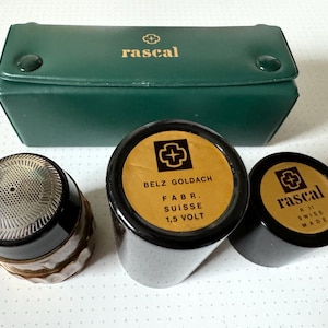Puede incluir: Tres contenedores redondos negros con etiquetas doradas. Las etiquetas tienen un símbolo de cruz y el texto "rascal", "BELZ GOLDACH FABR. SUISSE 1,5 VOLT" y "rascal 11 SWISS MADE". Los contenedores están sobre una superficie blanca. También se ve en la imagen una caja de cuero verde con la palabra "rascal" en letras doradas.