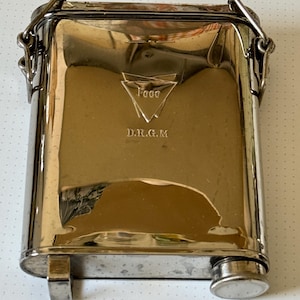 May include: A silver metal flask with a hinged lid and a chain handle. The flask has a small spout on the side and is engraved with the text "Foco D.R.G.M."