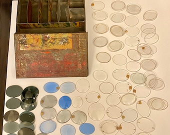 Könnte beinhalten: Eine geöffnete Metallbox mit Fächern, gefüllt mit verschiedenen Größen runder Glasuhrengläser. Die Box ist rot, gelb und grün mit einem Blumendesign. Auf einer weißen Oberfläche liegen auch mehrere Stapel von Uhrengläsern.