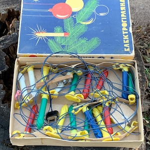 Op de afbeelding: Een vintage doos met kerstverlichting met een blauw-wit ontwerp. De doos bevat een lichtsnoer met gele, rode, blauwe en groene lampjes. De doos is gelabeld "Elektroprogramaal".