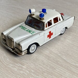 May include: White toy ambulance with a red cross and the word "AMBULANCE" printed in green on the side. The toy has blue flashing lights on the roof.