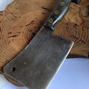 May include: A vintage cleaver with a dark handle featuring a horse head design. The handle has three brass rivets. The blade is rectangular with a hole near the corner. The cleaver rests on a wooden cutting board.