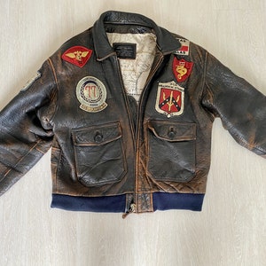 Puede incluir: Una chaqueta bomber de cuero marrón con varios parches, incluyendo un parche "77" con "Korea" escrito en él, un parche "U.S. Air Force" y un parche "Texas". La chaqueta tiene una cremallera y dos bolsillos con solapa.