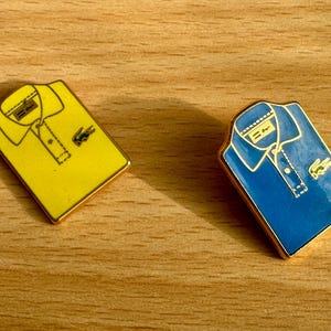 May include: Two enamel pins shaped like polo shirts. One is yellow, the other is blue, both with gold trim. Each pin features a collar, buttons, and a small green crocodile logo. The pins are on a wooden surface.
