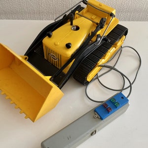 Puede incluir: Un bulldozer de juguete amarillo con una oruga negra y una pala grande amarilla. El bulldozer está conectado a un mando a distancia gris con botones azules y un cable negro.