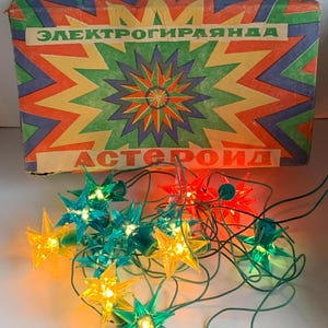 Könnte beinhalten: Eine vintage Lichterkette mit sternförmigen Glühbirnen in Grün, Gelb und Rot. Die Lichter befinden sich in ihrer Originalverpackung, die mit "Elektrogirlanda Asteroid" beschriftet ist.