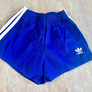 May include: Royal blue athletic shorts featuring an elasticized waistband, two white vertical stripes running down the side, and a white Adidas trefoil logo with the brand name printed on the lower right leg area of the garment.