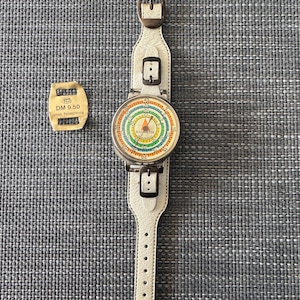 Puede incluir: Una pulsera de cuero blanco con un dispositivo circular plateado adjunto. El dispositivo tiene un dial colorido con la palabra "Kilómetro" impresa en él. El dispositivo también tiene una pequeña etiqueta adjunta que dice "DM 9.50" con las palabras "mit Freilauf" impresas debajo.