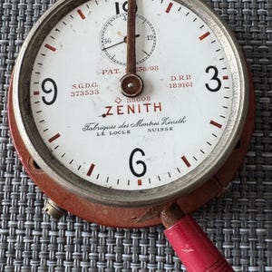 Op de afbeelding: Een vintage Zenith stopwatch met een witte wijzerplaat en rode en zilveren accenten. De stopwatch heeft een rode wijzer en een rode hendel. De wijzerplaat van de stopwatch heeft de tekst "ZENITH" en "Fabriques des Montres Zenith" in zwart. De tekst "S.G.D.G. 373533", "PAT. 536/08", "D.R.P. 189161" en "+35608" staat ook op de wijzerplaat gedrukt. De stopwatch is ingesteld op 6.