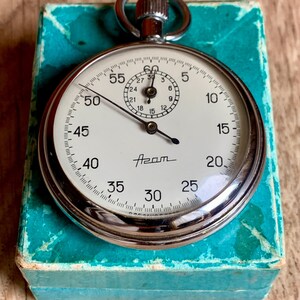 May include: A silver pocket watch with a white face and black hands. The watch has a black bezel with numbers from 5 to 60. The watch is in a blue box. The watch face says "Azam".