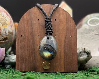 May include: A polished labradorite pendant with a dark grey and iridescent blue-gold flash, suspended from a black braided cord with a decorative knot. The pendant hangs against a smooth, arched wooden display stand with a small brass knob.