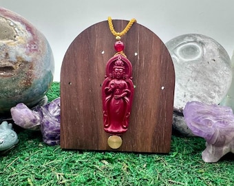 May include: A deep red carved pendant featuring a standing figure with an arched halo, suspended by a yellow cord with a red bead. The pendant is mounted on a dark brown, arched wooden display board with a small circular brass accent.
