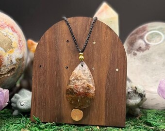 May include: A teardrop-shaped pendant made of clear, textured stone with brown, red, and white inclusions. The pendant hangs from a black braided cord featuring a small, round, gold-colored metal bead above the stone setting.