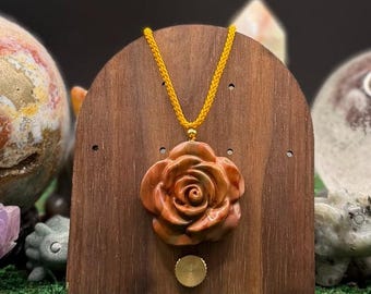 May include: A carved rose pendant made of brown and reddish-orange stone, suspended from a bright yellow braided cord. The pendant is displayed on a dark brown wooden stand with a smooth, arched top and a small circular metal accent below.