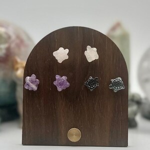 Puede incluir: Un expositor de madera con seis pares de pendientes de botón con forma de tortuga. Los pendientes están hechos de piedras blancas, moradas y negras. El expositor es marrón con un pequeño acento circular dorado en la parte inferior.