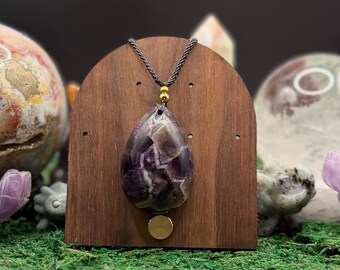 May include: A teardrop-shaped amethyst pendant with white quartz inclusions, suspended from a dark cord by a small gold-toned bead. The pendant is displayed on a curved, dark brown wooden stand with a circular gold-toned accent at the base.