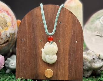 May include: A pale green carved stone fox pendant suspended on a light blue cord. A small, round, translucent red bead sits directly above the fox carving. The pendant is displayed on a dark brown, arched wooden stand with a circular knob.