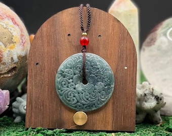 May include: A circular dark green jade pendant featuring intricate floral and swirling carvings, suspended by a brown braided cord with a single red bead and a small gold-toned accent, displayed on a curved wooden stand.