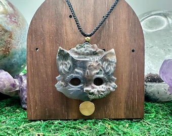 May include: A carved stone pendant in the shape of a cat head with hollow eyes, featuring mottled gray, white, and brown patterns. The pendant hangs from a black braided cord with a small gold-toned bead and a circular gold-toned charm.