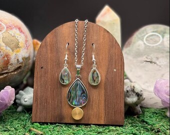 May include: A jewelry set featuring a silver chain necklace with a teardrop-shaped abalone shell pendant and a small gold-toned circular charm, accompanied by matching teardrop-shaped abalone shell earrings with silver-toned hooks.