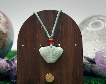 May include: A light green carved jade butterfly pendant suspended on a light green cord with a single small red bead accent. The pendant is displayed on a dark brown arched wooden stand with a small circular gold-toned metal knob.