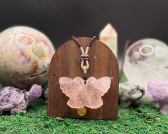 May include: A pale pink, translucent butterfly-shaped pendant carved from stone, suspended on a dark brown adjustable cord with a small gold-toned circular accent piece positioned directly below the butterfly on a wooden display stand.