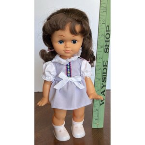 Peut inclure: Une poup&eacute;e vintage aux cheveux bruns, aux yeux bleus et v&ecirc;tue d'une robe lavande avec des manches blanches et un n&oelig;ud. La poup&eacute;e porte des chaussures et des chaussettes blanches. Une r&egrave;gle verte avec le texte "WAYSIDE SHOP" est &agrave; c&ocirc;t&eacute; de la poup&eacute;e.