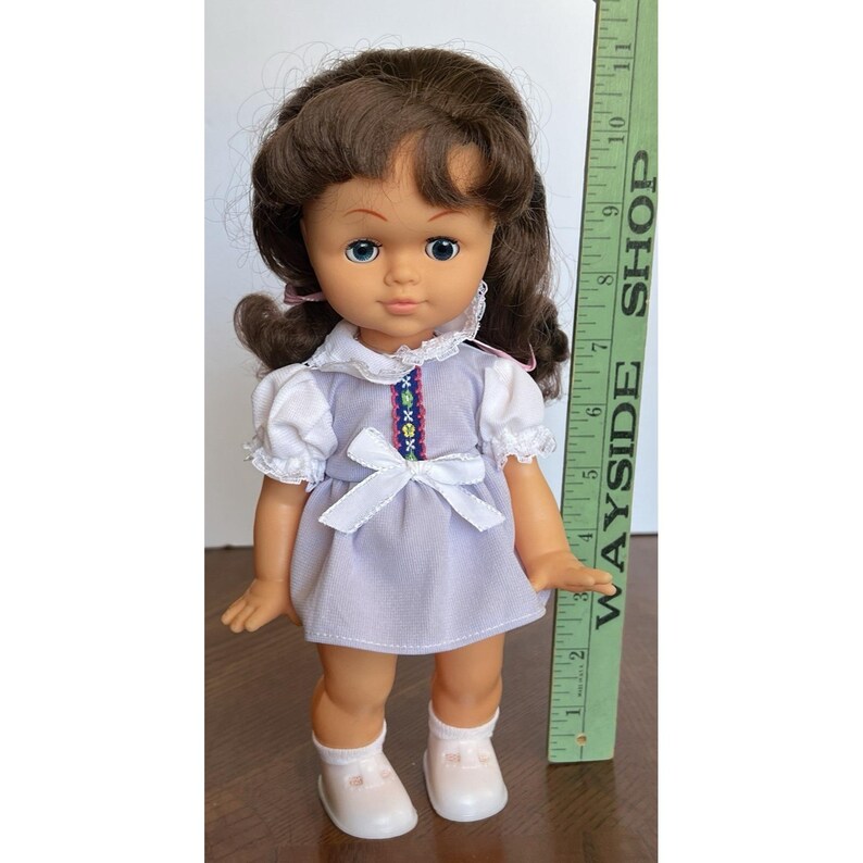 Peut inclure: Une poup&eacute;e vintage aux cheveux bruns, aux yeux bleus et v&ecirc;tue d'une robe lavande avec des manches blanches et un n&oelig;ud. La poup&eacute;e porte des chaussures et des chaussettes blanches. Une r&egrave;gle verte avec le texte "WAYSIDE SHOP" est &agrave; c&ocirc;t&eacute; de la poup&eacute;e.
