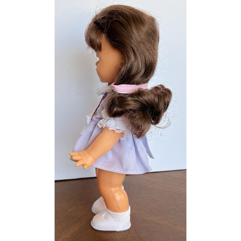 Peut inclure: Une poup&eacute;e vintage avec des cheveux bruns coiff&eacute;s en queue de cheval sur le c&ocirc;t&eacute;, v&ecirc;tue d'une robe violet clair avec une bordure en dentelle blanche. La poup&eacute;e porte des chaussures blanches et un ruban rose. La poup&eacute;e est sur une surface en bois.