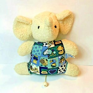 May include: A plush, cream-colored elephant toy with large, floppy ears. The elephant wears a patchwork vest with blue, green, and white squares featuring dog-themed illustrations. A small, wooden bead hangs from a string at the bottom.