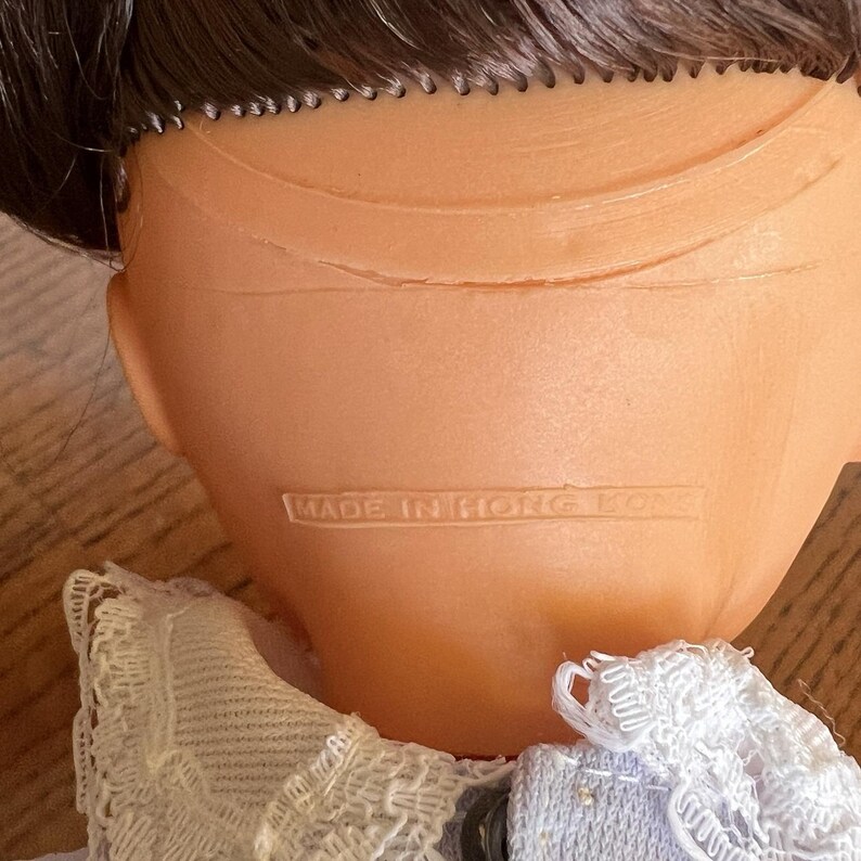 Peut inclure: T&ecirc;te de poup&eacute;e vintage, vue de dos, avec des cheveux bruns et une construction en plastique. Le texte "MADE IN HONG KONG" est imprim&eacute; au dos de la t&ecirc;te. La poup&eacute;e porte un col en dentelle blanche.