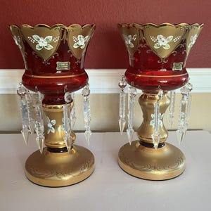 May include: Pair of ruby red glass candle holders with gold accents. Each holder features a scalloped rim with gold trim and white floral designs. Clear crystal prisms hang from the base, adding sparkle. The base is gold with decorative swirls.