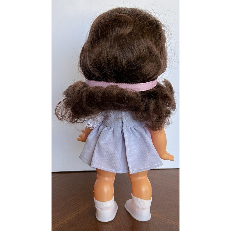 Peut inclure: Vue arri&egrave;re d'une poup&eacute;e vintage avec des cheveux bruns boucl&eacute;s et un ruban rose clair. La poup&eacute;e porte une robe bleu clair et des chaussettes blanches avec des chaussures blanches. La peau de la poup&eacute;e est de couleur p&ecirc;che claire.