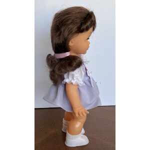 Peut inclure: Poup&eacute;e vintage aux cheveux bruns attach&eacute;s en queue de cheval, v&ecirc;tue d'une robe violet clair avec une bordure en dentelle blanche. La poup&eacute;e porte des chaussures blanches et un ruban rose. La poup&eacute;e est sur une surface en bois.